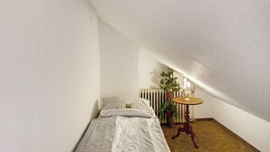 A small, single-bed room with a radiator, a table with a lamp, and a potted plant.