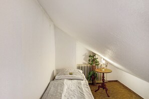 A small, single-bed room with a radiator, a table with a lamp, and a potted plant.