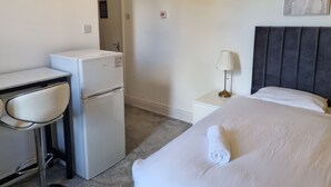 Hotellihuone, jossa on sänky, työpöytä, jossa on jääkaappi, tuoli, yöpöytä, jossa on lamppu, ja seinälle kiinnitetty taideteos.