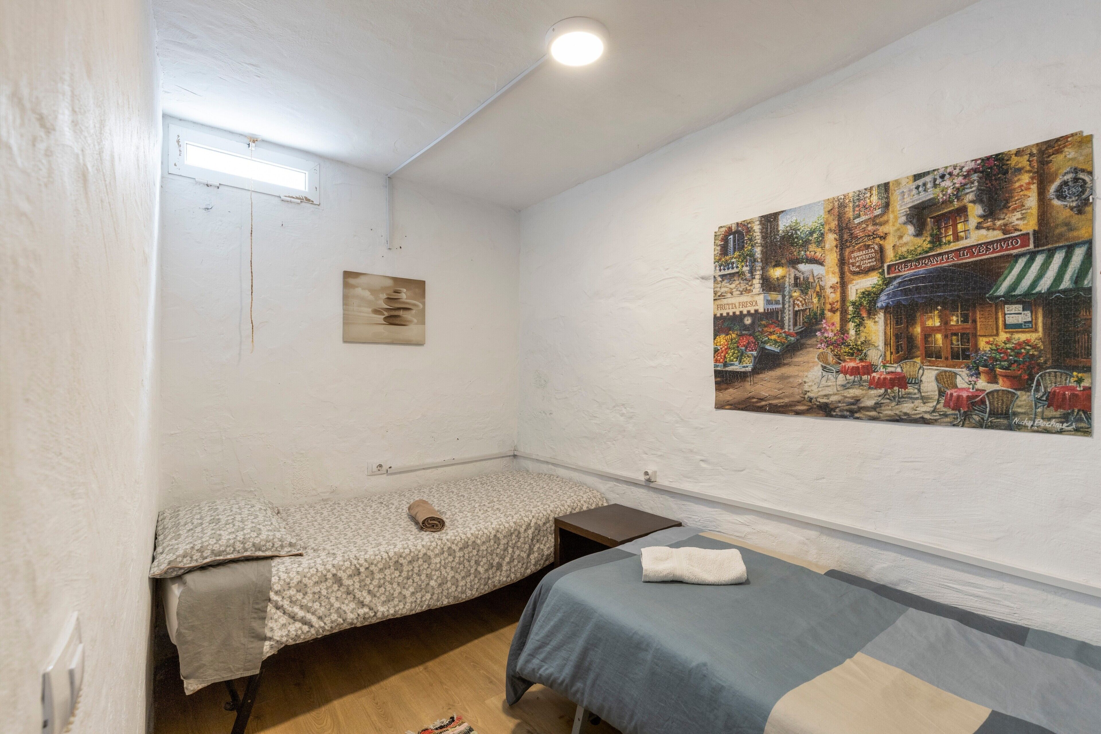 A room with two beds, a small table, a wall-mounted painting, and a ceiling light.