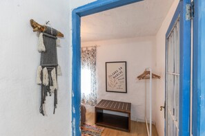 Um quarto com porta azul, um painel de macramé fixado na parede, uma moldura com uma frase, um banco de madeira e um cabide suspenso.