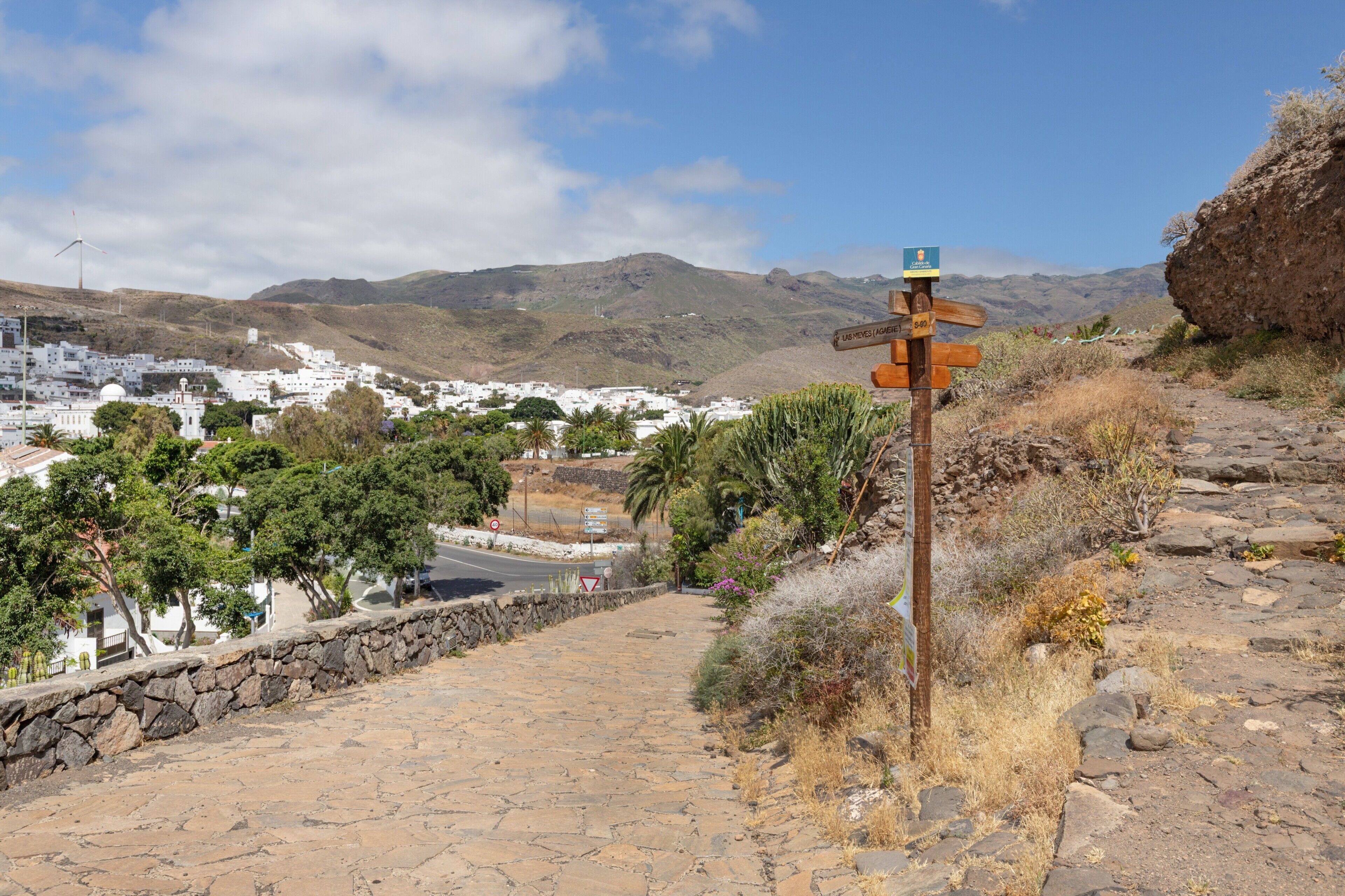 A stone pathway leading to a signpost with multiple directions, surrounded by dry vegetation and a distant town.