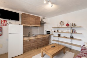 Een kleine keuken met houten kastjes, een witte koelkast, een magnetron en een koffiezetapparaat. Er staat een houten tafel met een afstandsbediening erop.