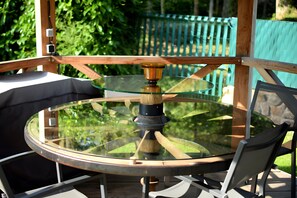 Une table ronde en verre, dotée d’un pied central en bois, soutenue par des pieds en métal, est placée à l’extérieur, sur une terrasse en bois, avec une clôture verte en arrière-plan.
