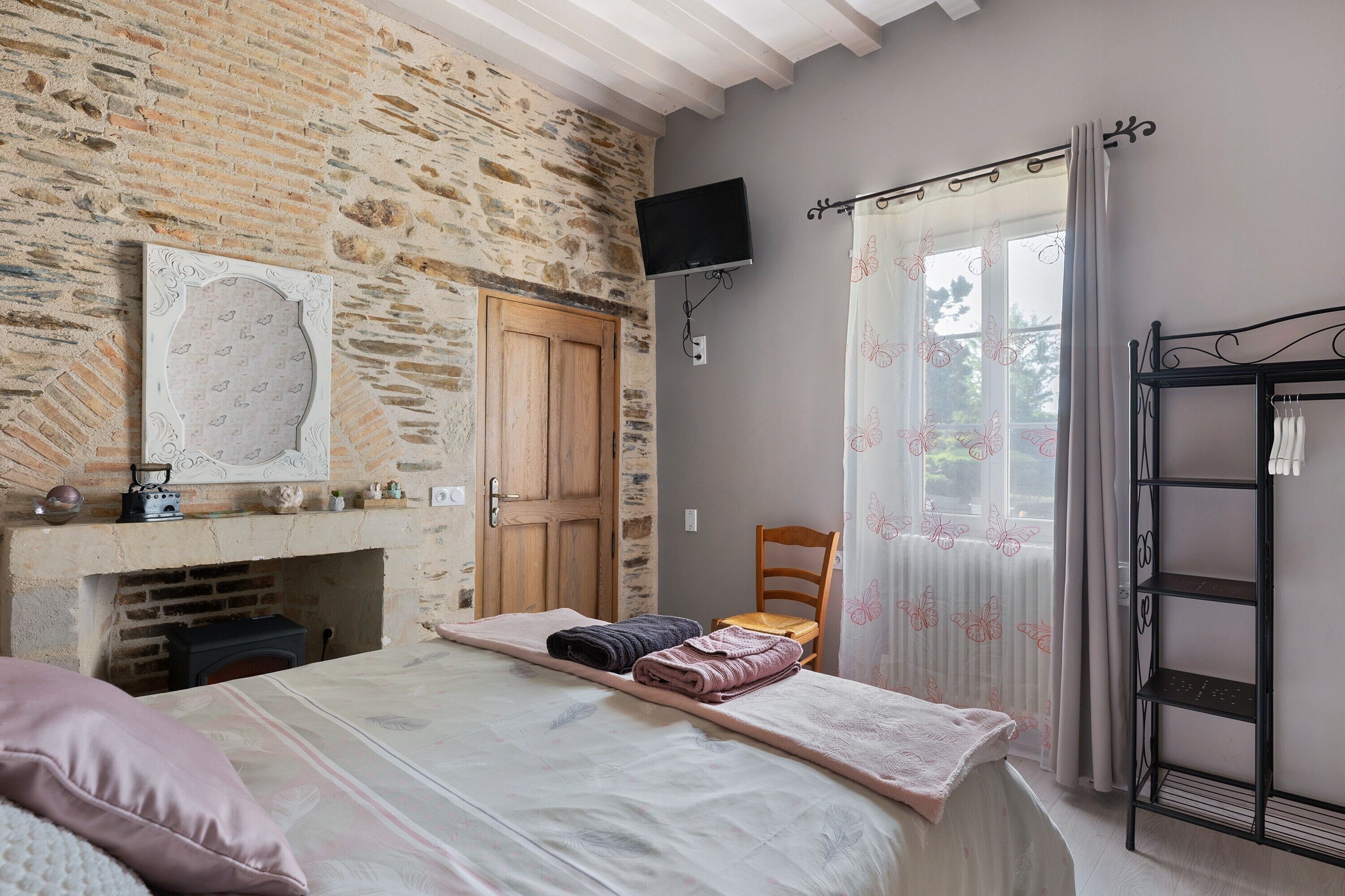 A room with a stone wall, a wooden door, a fireplace, a bed with a blanket, a chair, a TV, and a window with curtains.