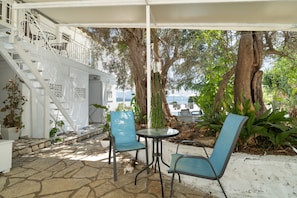 A covered outdoor seating area with stone flooring, two blue chairs, a small table, and a staircase leading to an upper level. - Room 'Ground Floor' with Sea View, Shared Pool and Wi-Fi (Nikiana)
