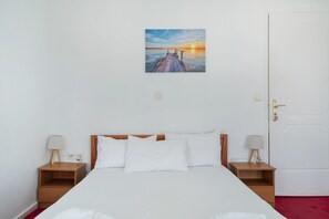 Ein ordentlich eingerichtetes Hotelzimmer mit einem Bett, zwei Nachttischen mit Lampen, einer weißen Tür und einem Bild von einem Sonnenuntergang über dem Wasser an der Wand.