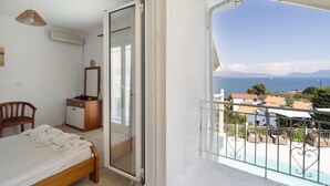 Een slaapkamer met een bed, een stoel, een dressoir en een schuifdeur naar een balkon met uitzicht op zee en gebouwen.