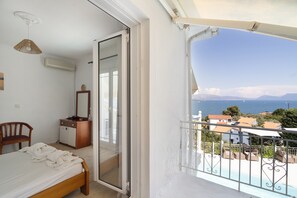 A bedroom with a bed, a chair, a dresser, and a sliding glass door leading to a balcony with a view of the sea and buildings. - Room '1st Floor' with Sea View, Shared Pool and Wi-Fi (Nikiana)