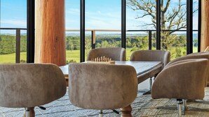 Una mesa de madera con un tablero de ajedrez, cuatro sillas y ventanas amplias que dan a un paisaje verde.