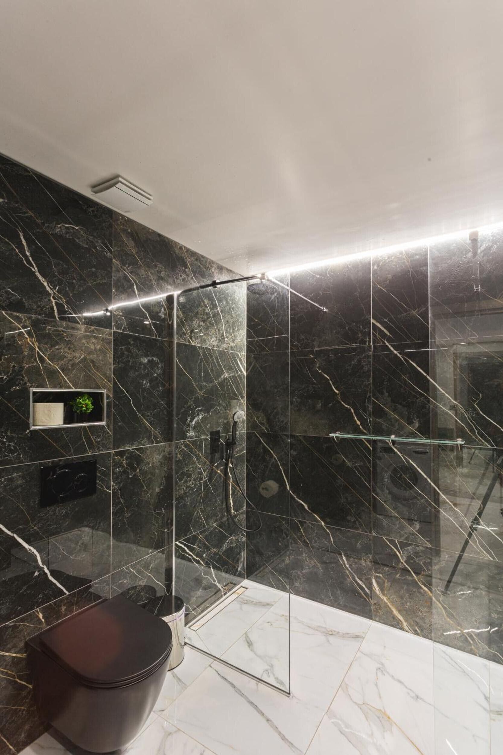 Un baño moderno con ducha envuelta en vidrio, paredes de mármol y un inodoro de tonos oscuros.