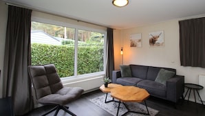 Ein modernes Wohnzimmer mit einer Couch, einem Sessel, einem Couchtisch und Blick auf einen Garten durch ein großes Fenster.