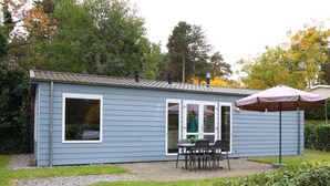 Ein kleines, eingeschossiges Gebäude mit einer Terrasse, die einen Tisch und Stühle sowie eine überdachte Sitzgelegenheit bietet, und einem Kiesweg, der zum Eingang führt.