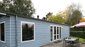 Ein kleines, eingeschossiges Gebäude mit einer Terrasse, die einen Tisch und Stühle sowie eine überdachte Sitzgelegenheit und einen kleinen Garten mit einer Hecke bietet.