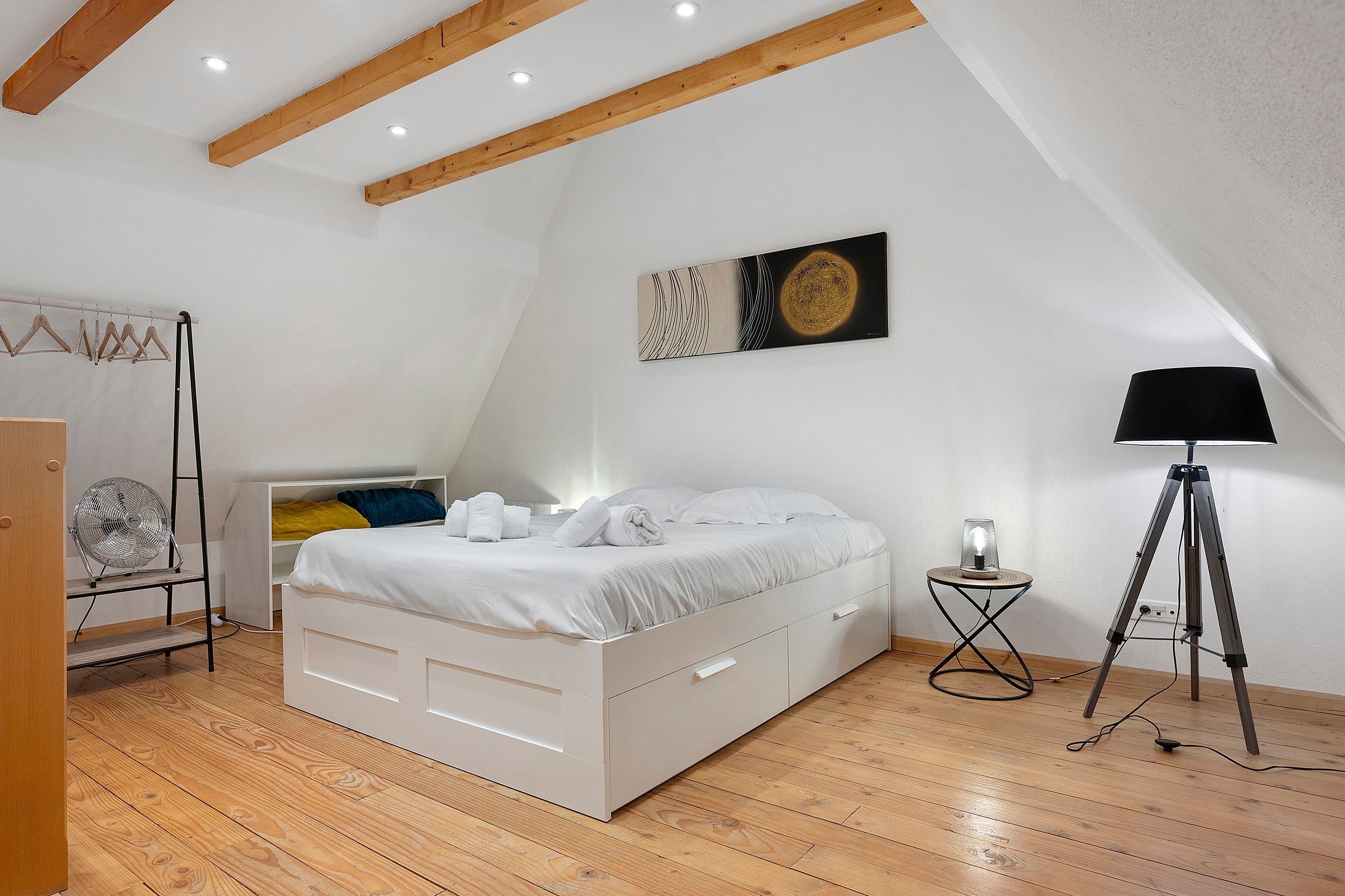 Une chambre avec un lit blanc, des poutres en bois, un ventilateur de plafond, une lampe sur pied et une œuvre d’art accrochée au mur.