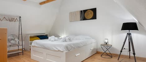 Een slaapkamer met een wit bed, houten balken, een plafondventilator, een vloerlamp en een kunstwerk aan de muur.