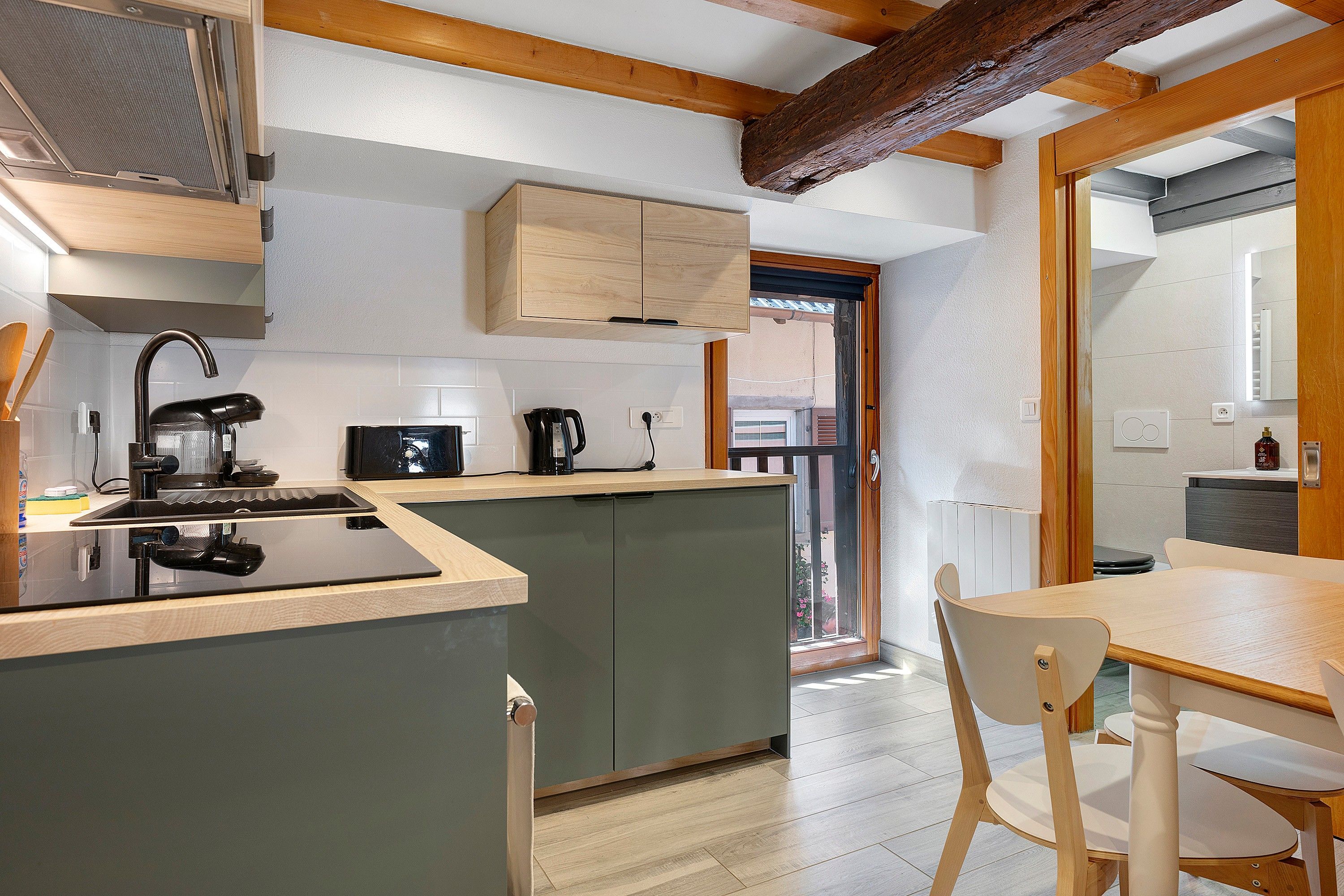 Cocina compacta con isla integrada, zona de comedor con mesa y sillas de madera, y vista a través de un ventanal abierto.