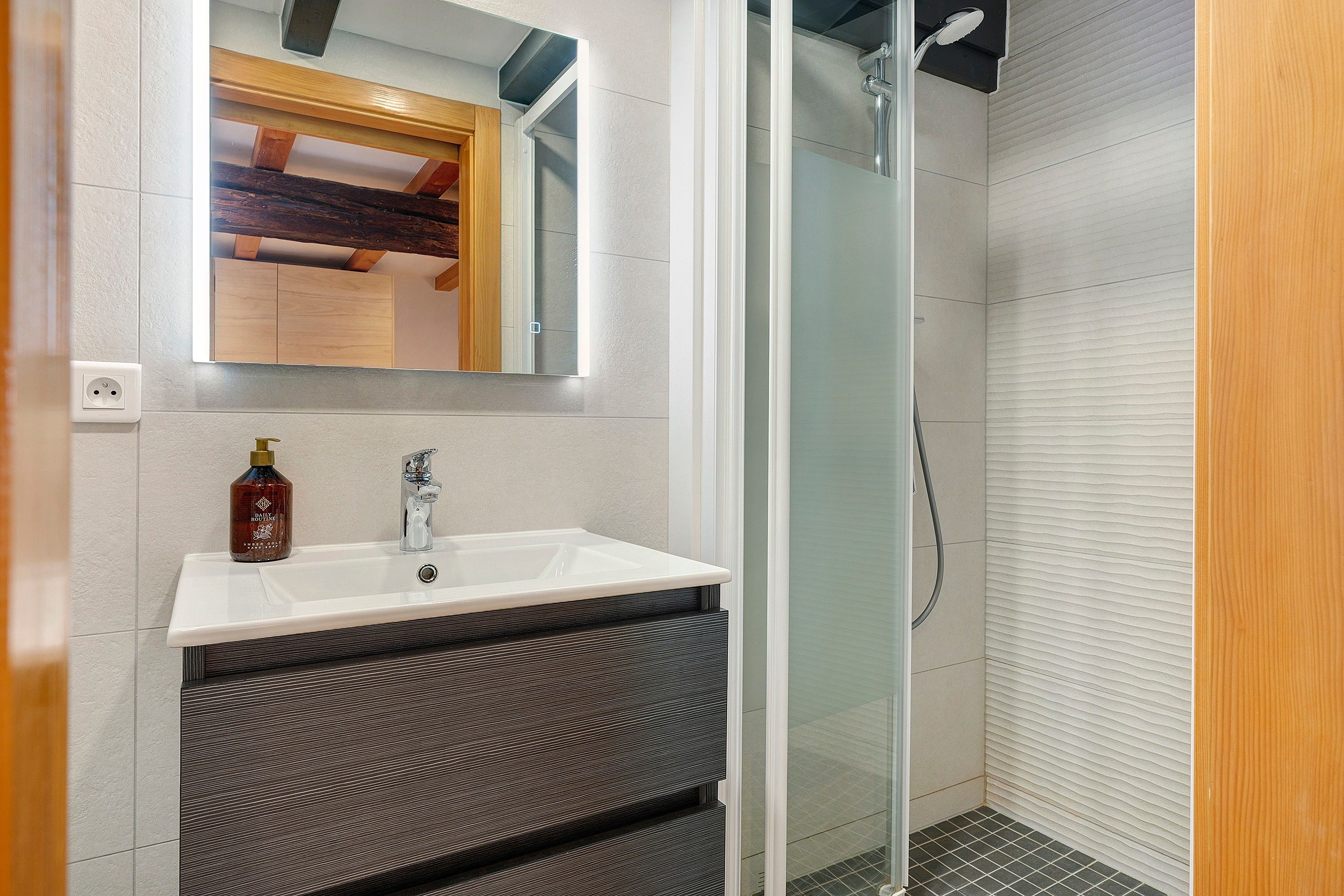 Une salle de bain avec un lavabo blanc, un miroir, un flacon de savon liquide et un espace douche.
