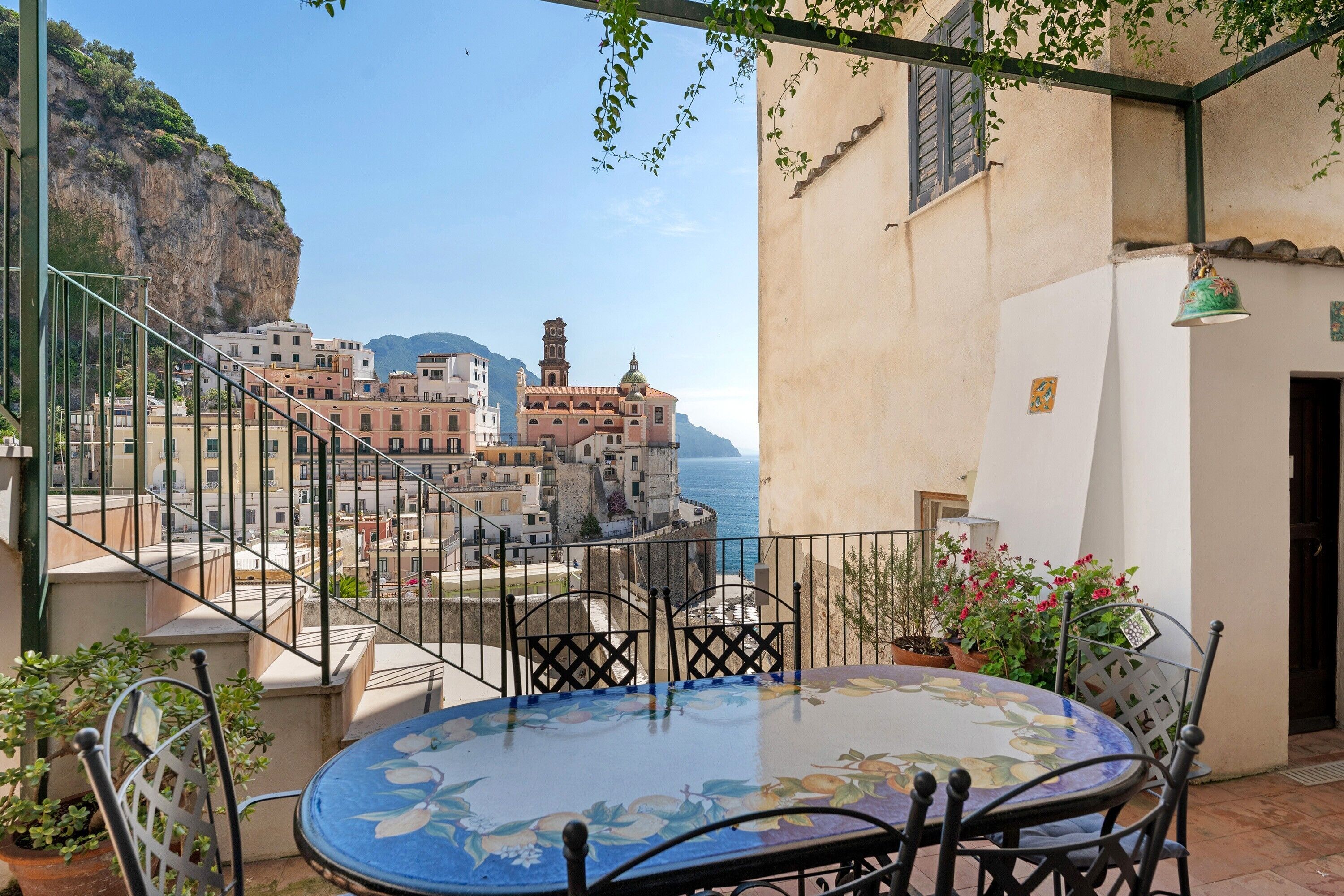 A balcony with a table and chairs, overlooking a coastal town with historic buildings and a clear view of the sea.