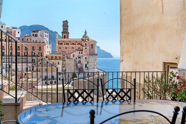 Un balcon avec une table, une vue sur une ville côtière avec ses bâtiments et son église, et un ciel bleu dégagé.
