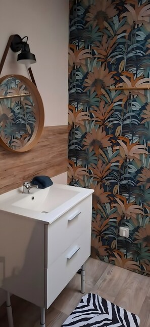 A bathroom with a white sink, a mirror with a wooden frame, a wall-mounted light, and a patterned wallpaper. - Studio Apartment 'Lisaline 2' with Wi-Fi (Saint-Dyé-sur-Loire)