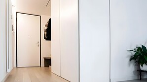 Un couloir avec un plancher en bois, des murs blancs, une porte, un petit banc et une plante en pot.