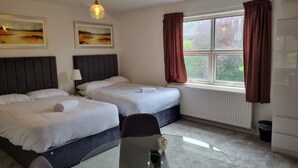 Ein Hotelzimmer mit zwei Betten, einem runden Glastisch, einem Sessel, einer Lampe, einem Fenster mit Vorhängen und zwei Bildern an der Wand.