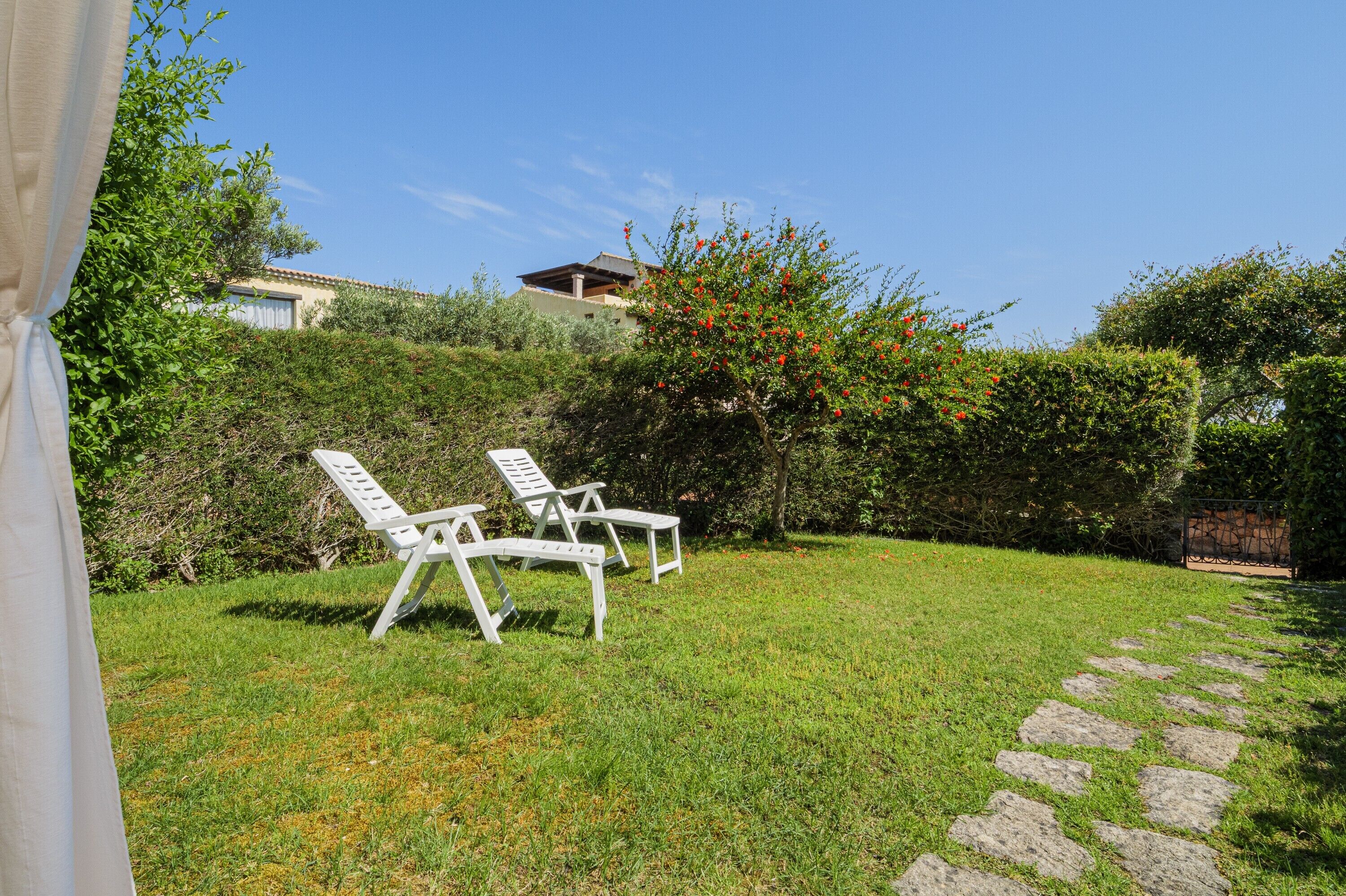 Ein Garten mit Rasen, zwei weißen Liegestühlen, einem Kiesweg und einem Haus im Hintergrund.