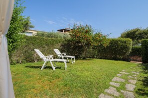 Un patio trasero con césped, dos sillas reclinables blancas, un camino de piedra y una casa al fondo.