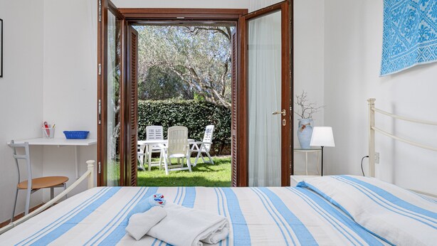 Quarto com cama, janela, escrivaninha, cadeira, mesa e vista para um jardim.
