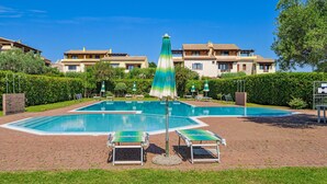 Piscina com espreguiçadeiras e guarda-sóis, cercada por sebes e edifícios.