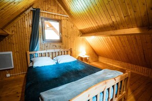 Una habitación de madera con una cama, un ventanal con cortinas, una mesita de noche con una lámpara y un radiador.