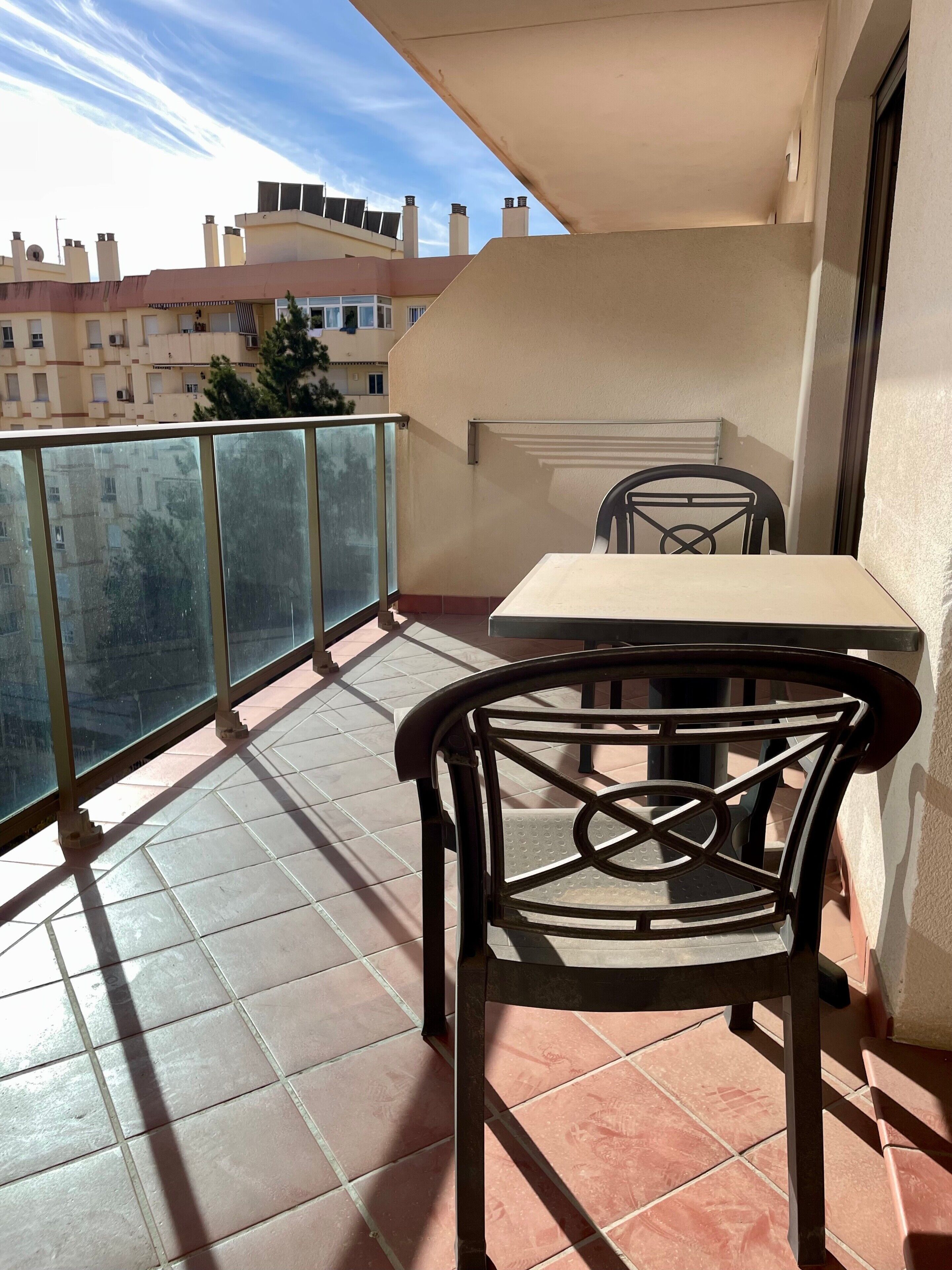 A balcony with a table and chair, a tiled floor, and a view of buildings in the background.