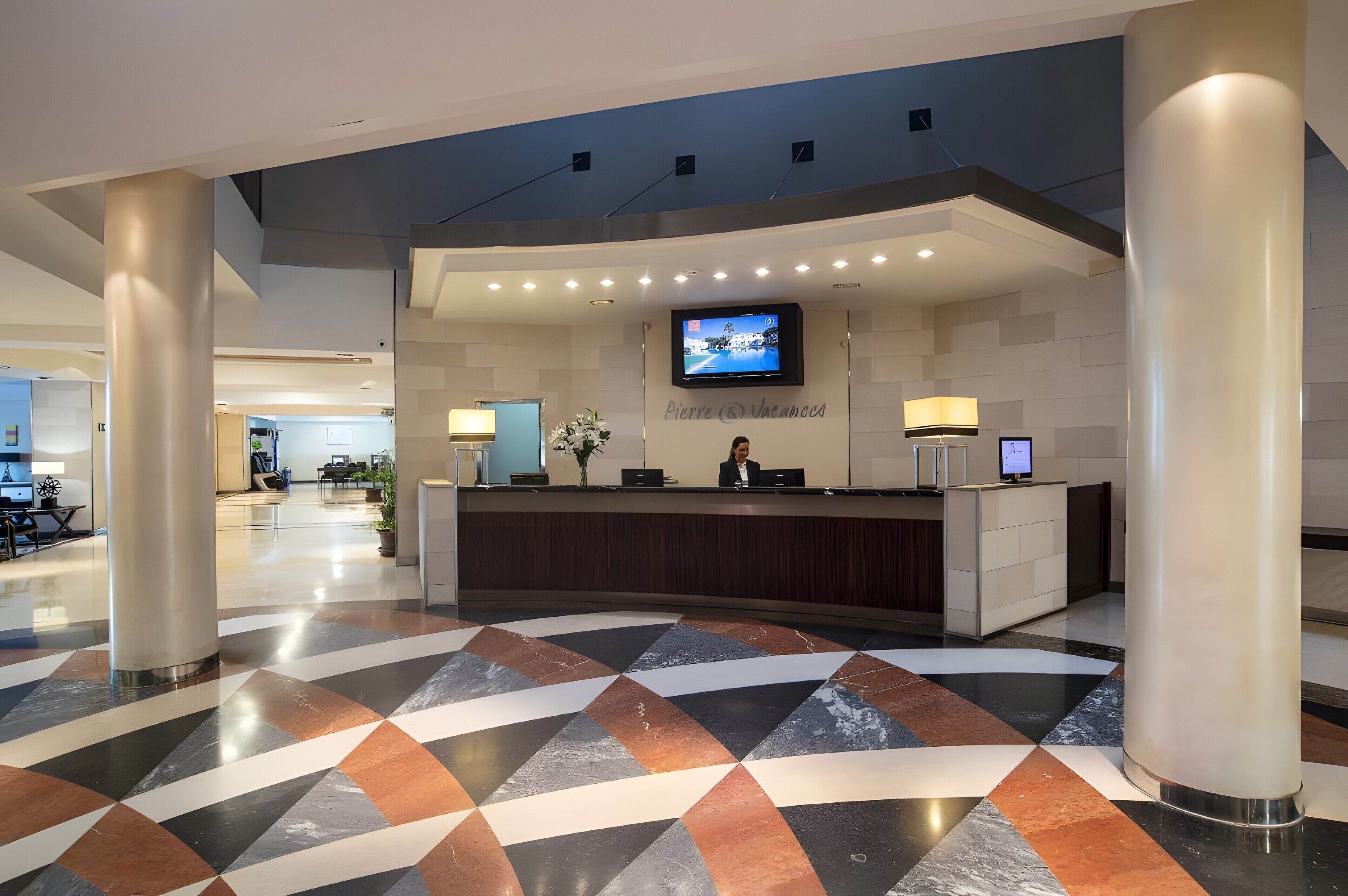 Zona de recepción de un hotel con diseño moderno, que incluye un mostrador de recepción, una pantalla de televisión y un suelo con estampado.