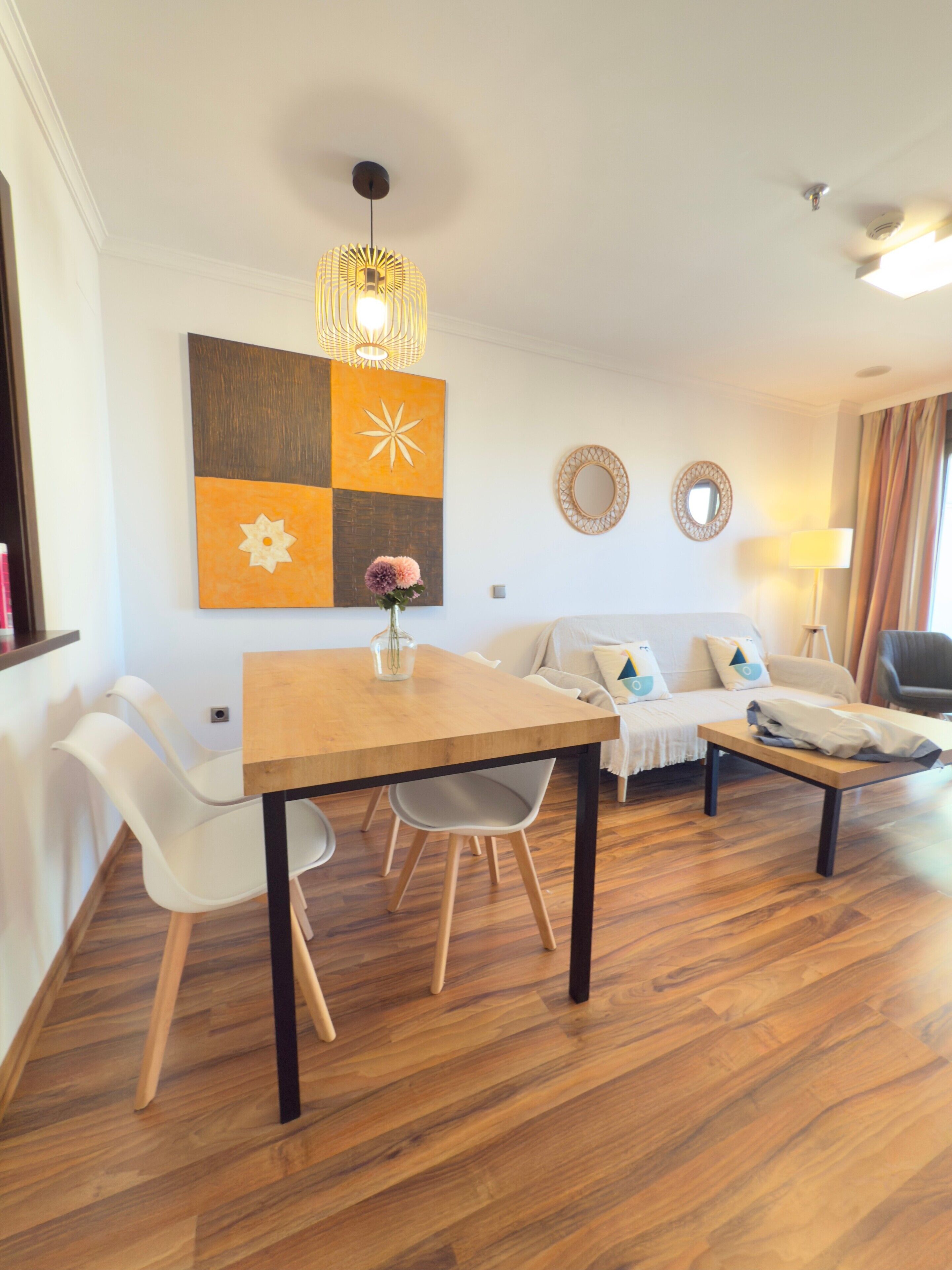 Una sala de estar moderna con una mesa de comedor de madera, sillas blancas, un sofá con cojines y un elemento decorativo en la pared.
