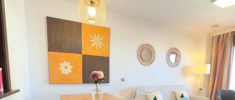 Una sala de estar moderna con una mesa de comedor de madera, sillas blancas, un sofá con cojines y un elemento decorativo en la pared.