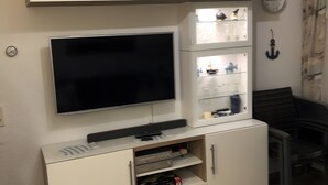 Uma sala de estar com televisão de tela plana presa a um móvel, um relógio na parede e uma estante com diversos objetos.
