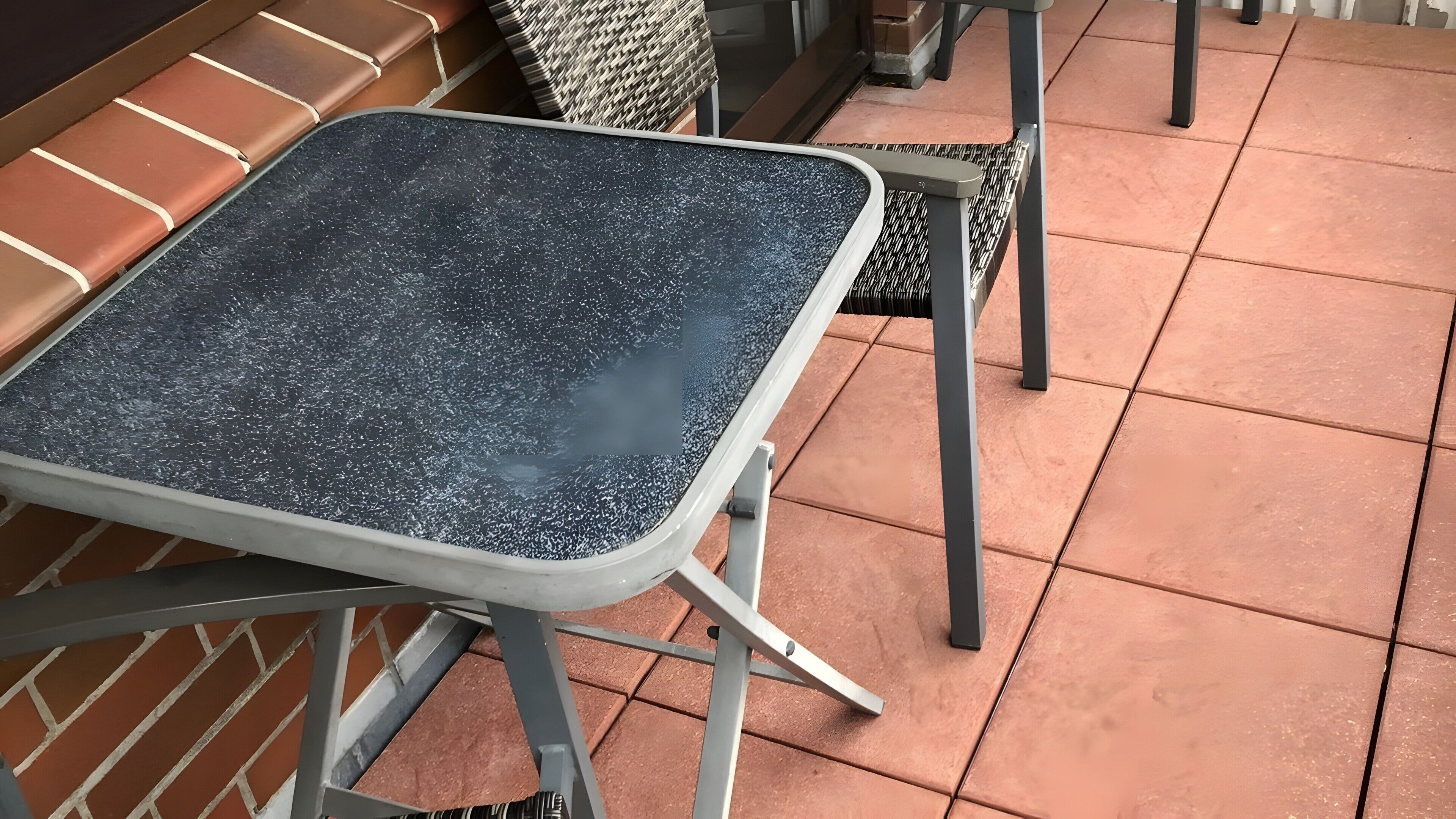 Ein kleiner Tisch im Freien mit Metallgestell und einer schwarz gefleckten Tischplatte, platziert auf einer gefliesten Terrasse mit einer Backsteinmauer im Hintergrund.