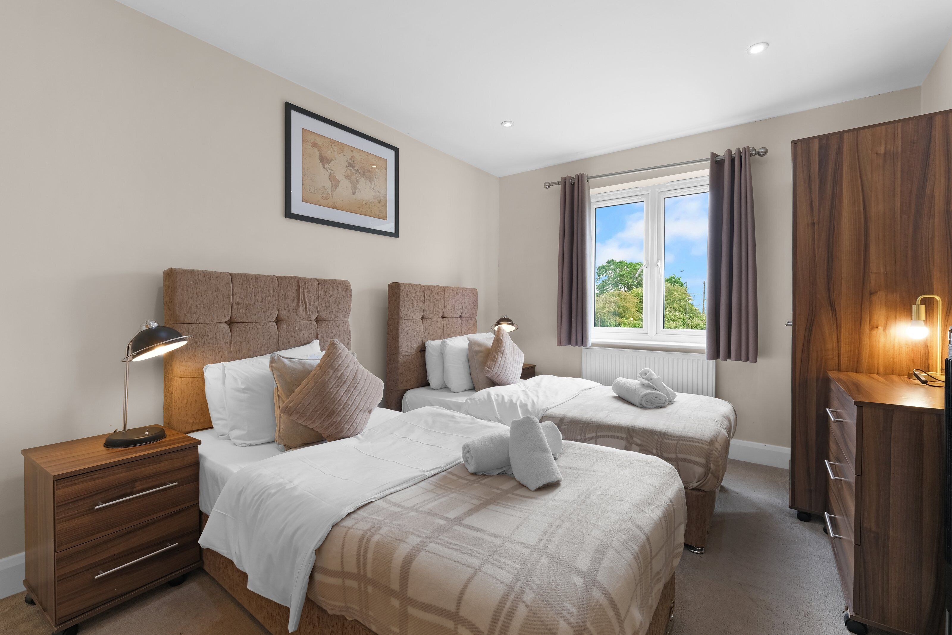 Quarto de hotel com duas camas, uma mesinha de cabeceira de madeira, cômoda e janela com cortinas.