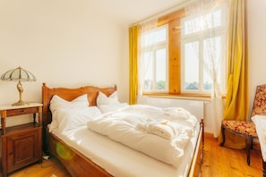 Un lit bien fait, avec du linge de lit blanc, une table de chevet en bois surmontée d’une lampe, une chaise agrémentée d’un coussin à motifs floraux et une fenêtre ornée de rideaux jaunes.