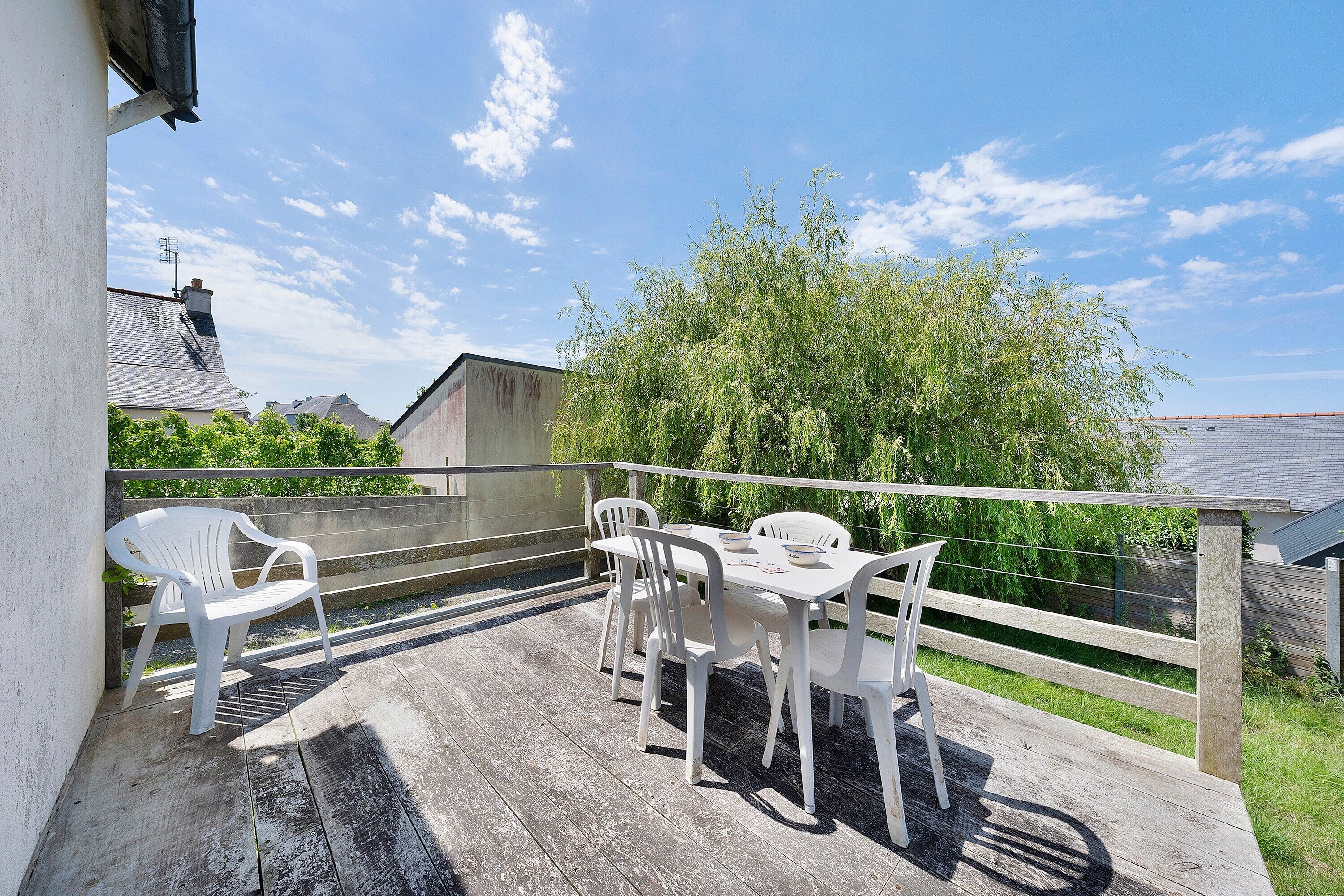 Eine Holzterrasse mit Tisch und Stühlen, einem Geländer sowie Blick auf Bäume und Gebäude.