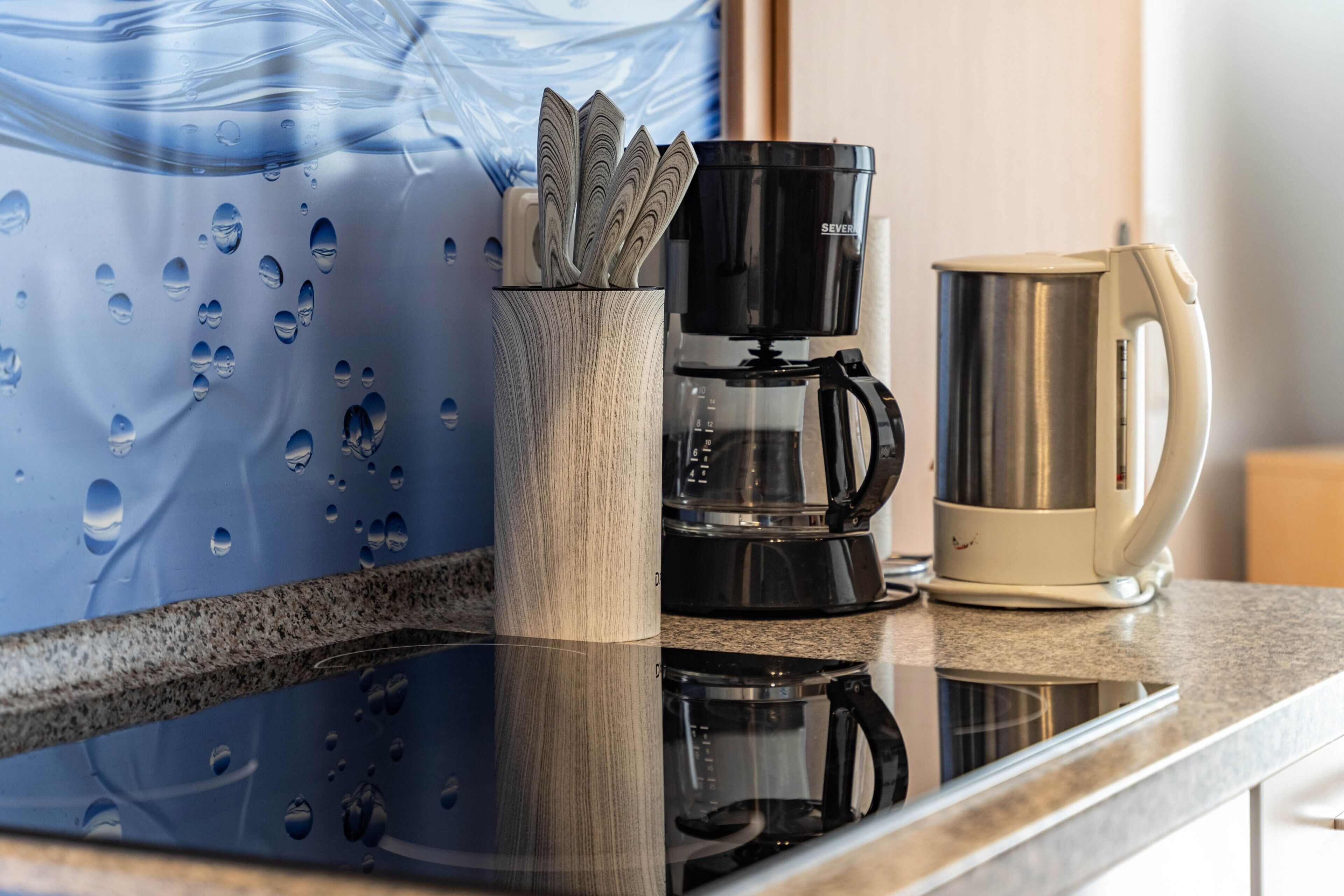 A kitchen counter with a coffee maker, a kettle, and a glass container with utensils.