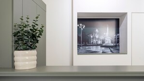 Uma planta em vaso sobre uma prateleira, uma fotografia a preto e branco emoldurada de um edifício, e uma parede branca.