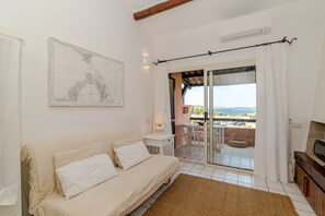 Quarto com sofá, mesa, cadeira, televisão e vista para o mar através de uma porta de vidro.
