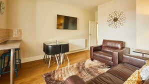 Una sala de estar con un sofá de cuero marrón, una mesa de comedor con sillas, un televisor montado en la pared y un adorno decorativo en la pared.