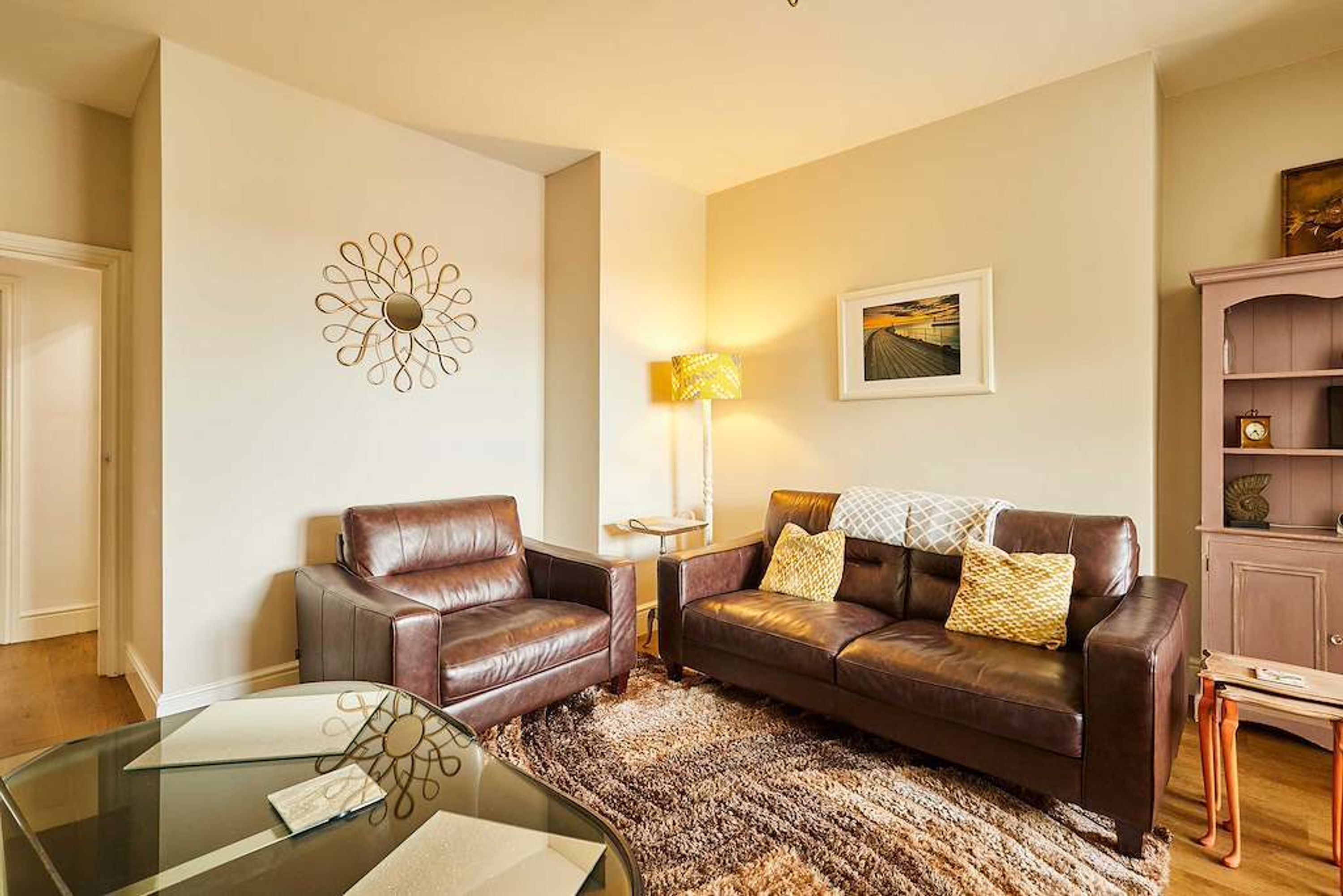 A living room with a brown leather sofa, a glass coffee table, a patterned rug, a wall art piece, and a lamp.