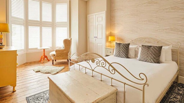 Un dormitorio con cama, una silla, una mesita de noche, una lámpara y una ventana con persianas.
