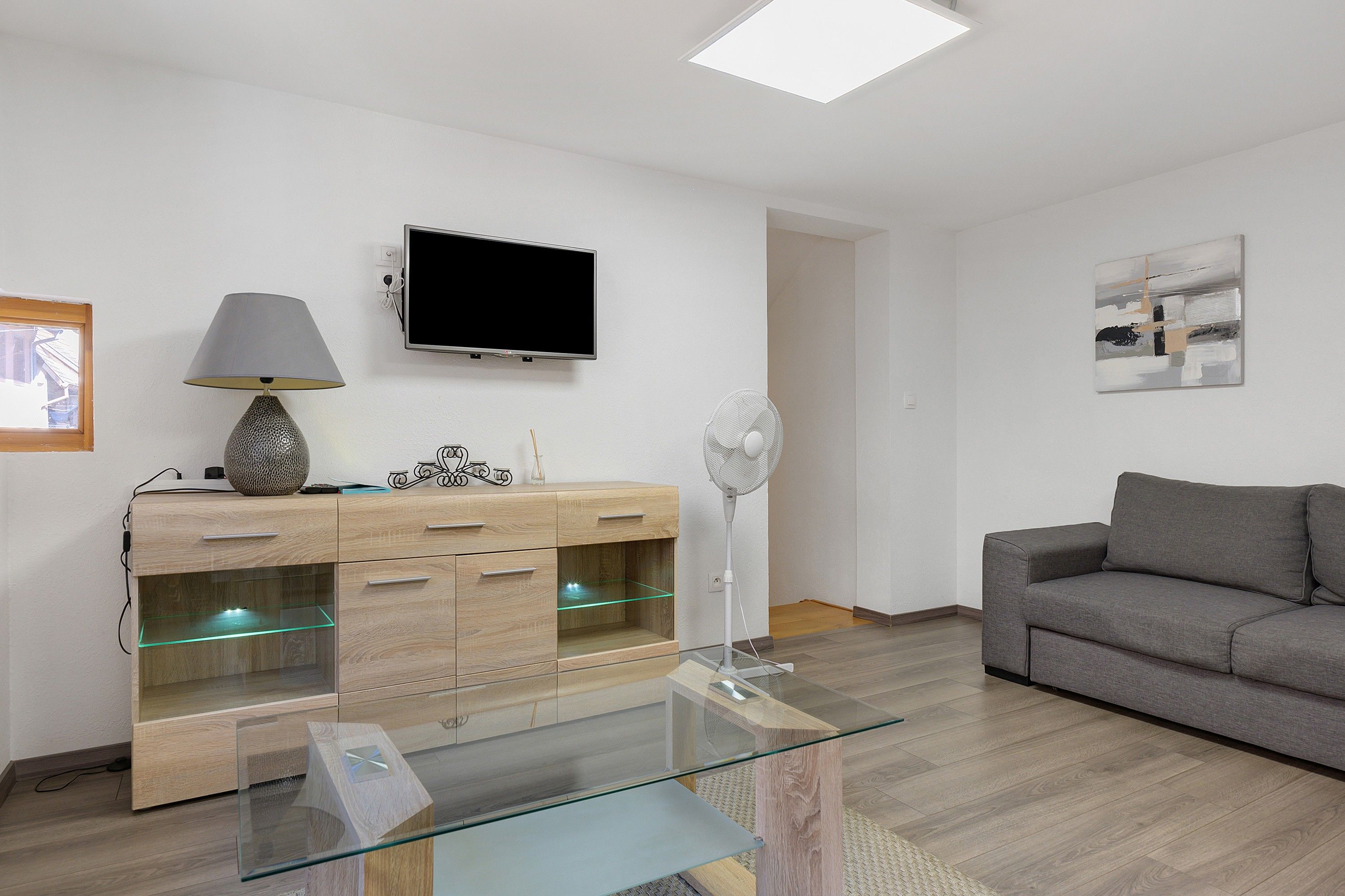 Un salon moderne avec une table basse en verre, un canapé gris, un meuble en bois avec des tiroirs, un ventilateur sur pied et un téléviseur à écran plat fixé au mur.