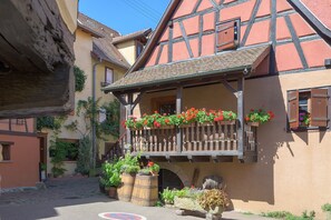 A traditional half-timbered building with a balcony adorned with flowers, a cobblestone street, and a stone barrel. - Le Zen d'Eguisheim – A Timeless Escape (Eguisheim)