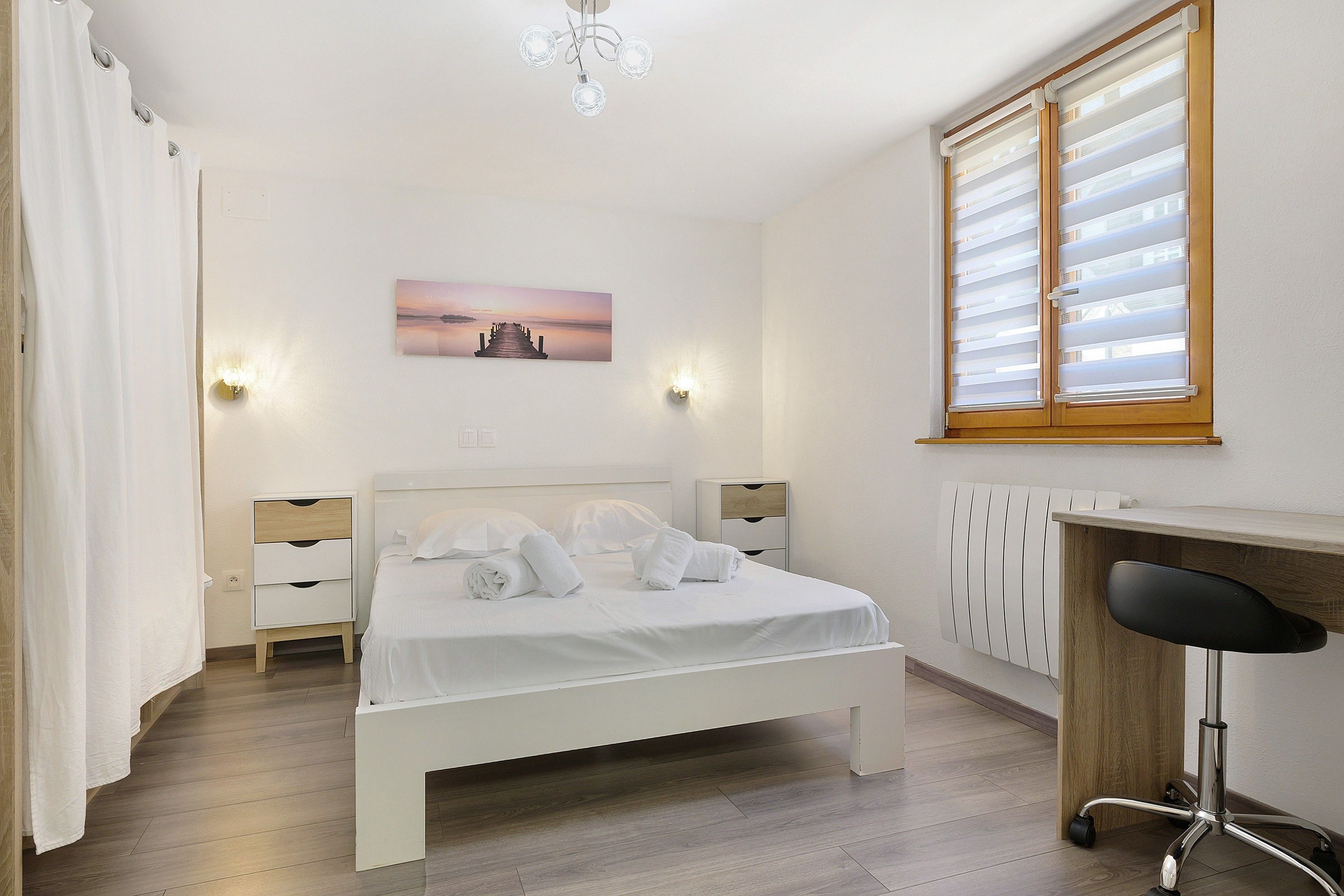 A hotel room with a bed, bedside tables, a desk, a chair, a radiator, and a window with blinds.